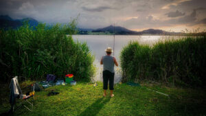 Ein Angler steht mit seiner Angel am Ufer eines Sees. Der Zugang zum See wird an beiden Seiten von einem dichten Schilfgürtel begrenzt. Das aufziehende Unwetter am Horizont scheint ihm nicht zu stören.