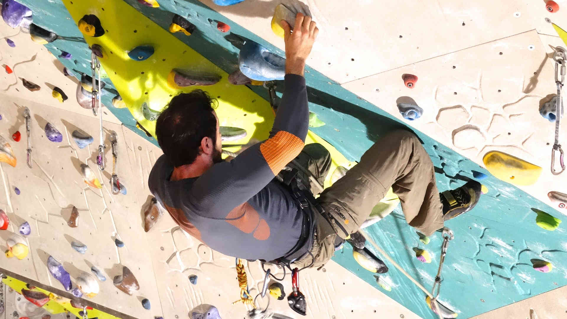 Das Bild zeigt einen Kletterer beim Bouldern. Es findet in einer Kletterhalle an einer speziellen Kletterwand statt. Er ist mit einem Seil gesichert.