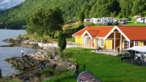 Am felsigen Ufer eines See stehen Zelte und Caravans. Zentrale bildet ein gelbes Holzhaus. Im Hintergrund gibt es viel Wald.