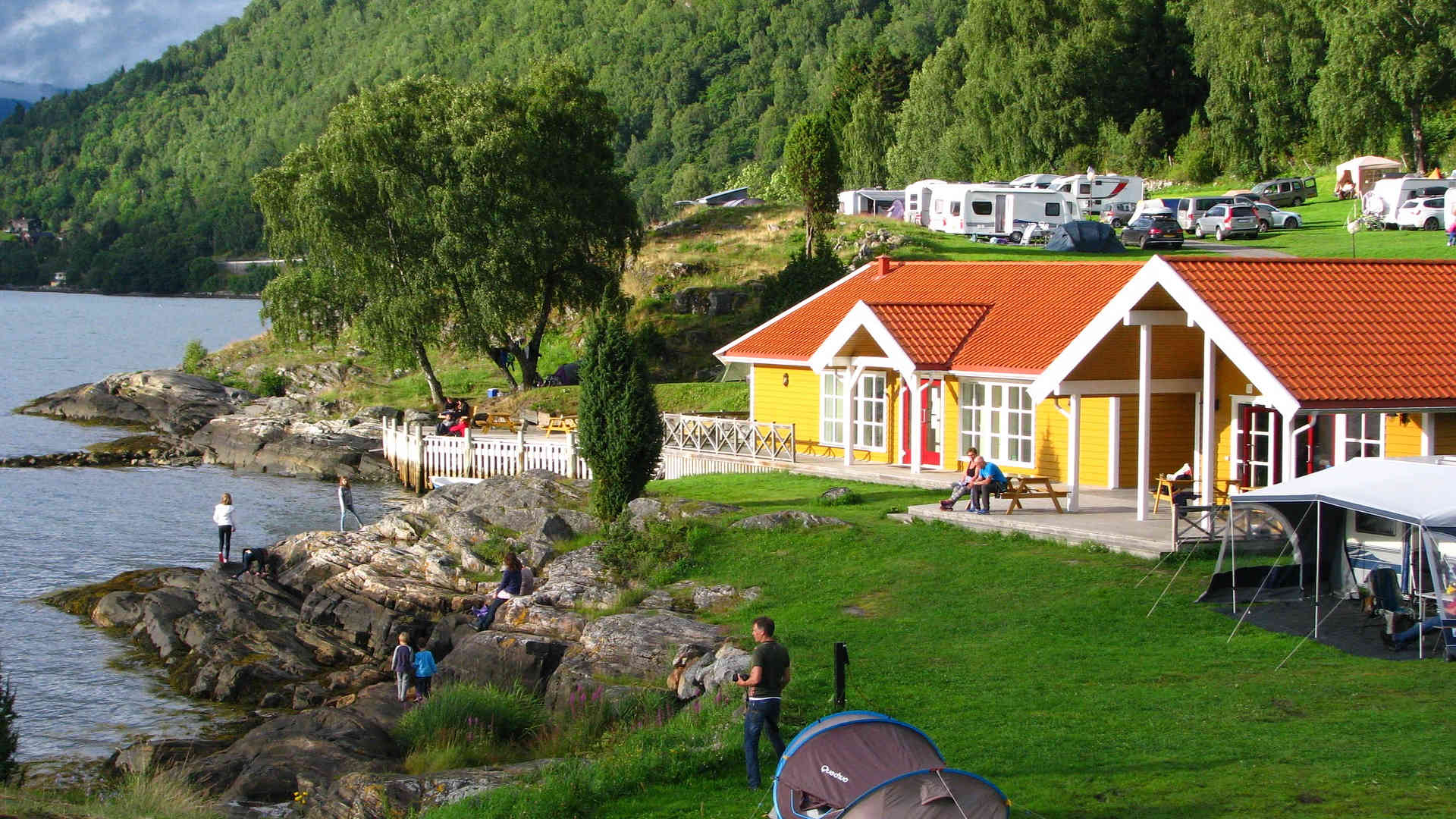 Am felsigen Ufer eines See stehen Zelte und Caravans. Zentrale bildet ein gelbes Holzhaus. Im Hintergrund gibt es viel Wald.