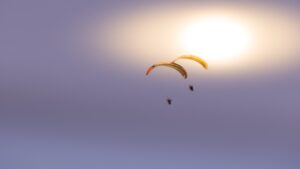 Zwei Motorschirm-Piloten fliegen dicht beieinander im Formationsflug. Aus der Sicht des Beobachters passieren sie gerade die durch Hochnebel leicht bedeckte Sonne.