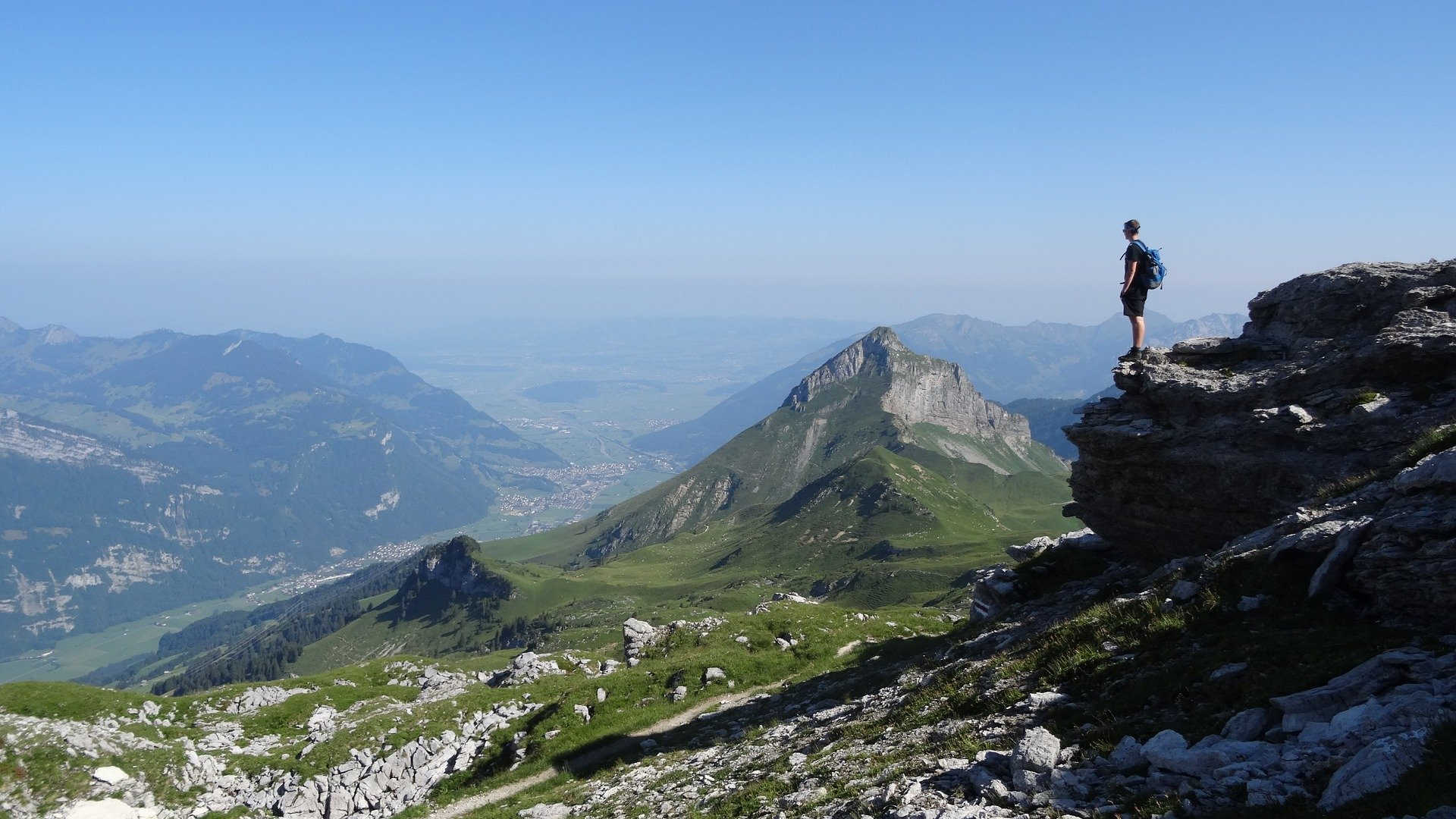 Ein Wanderer steht hoch oben in den Bergen auf einem Felsen. Er genießt die Aussicht ins Tal und auf die anderen Berggipfel. Die Fernsicht ist heute hervorragend.