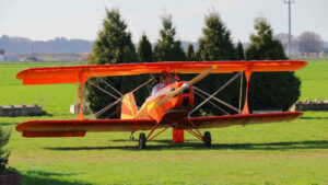 Ein Doppeldecker steht auf einer grünen Wiese. Seine Lackierung ist auffällig orange mit gelben Akzenten. Er scheint abflugbereit und wartet nur noch auf seinen Piloten.