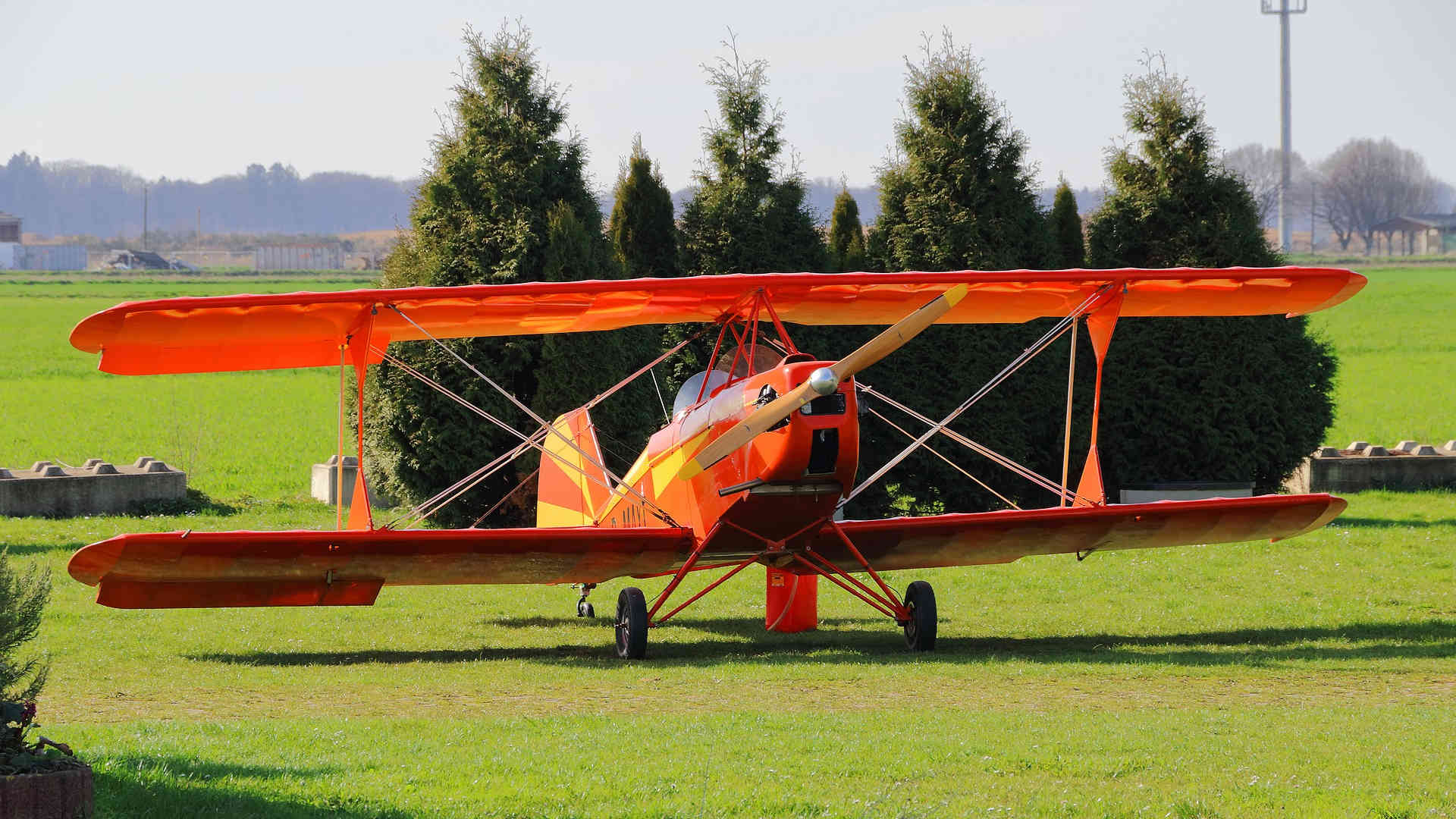 Ein Doppeldecker steht auf einer grünen Wiese. Seine Lackierung ist auffällig orange mit gelben Akzenten. Er scheint abflugbereit und wartet nur noch auf seinen Piloten.