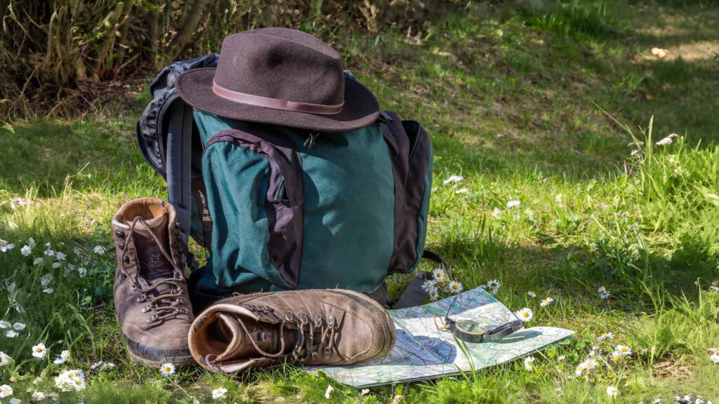 Auf einem Wanderrucksack liegt ein Filzhut. Davor stehen auf der Gänsblümchenwiese Wanderschuhe. Eine Wanderkarte und ein Kompass ergänzen das Ensemle.