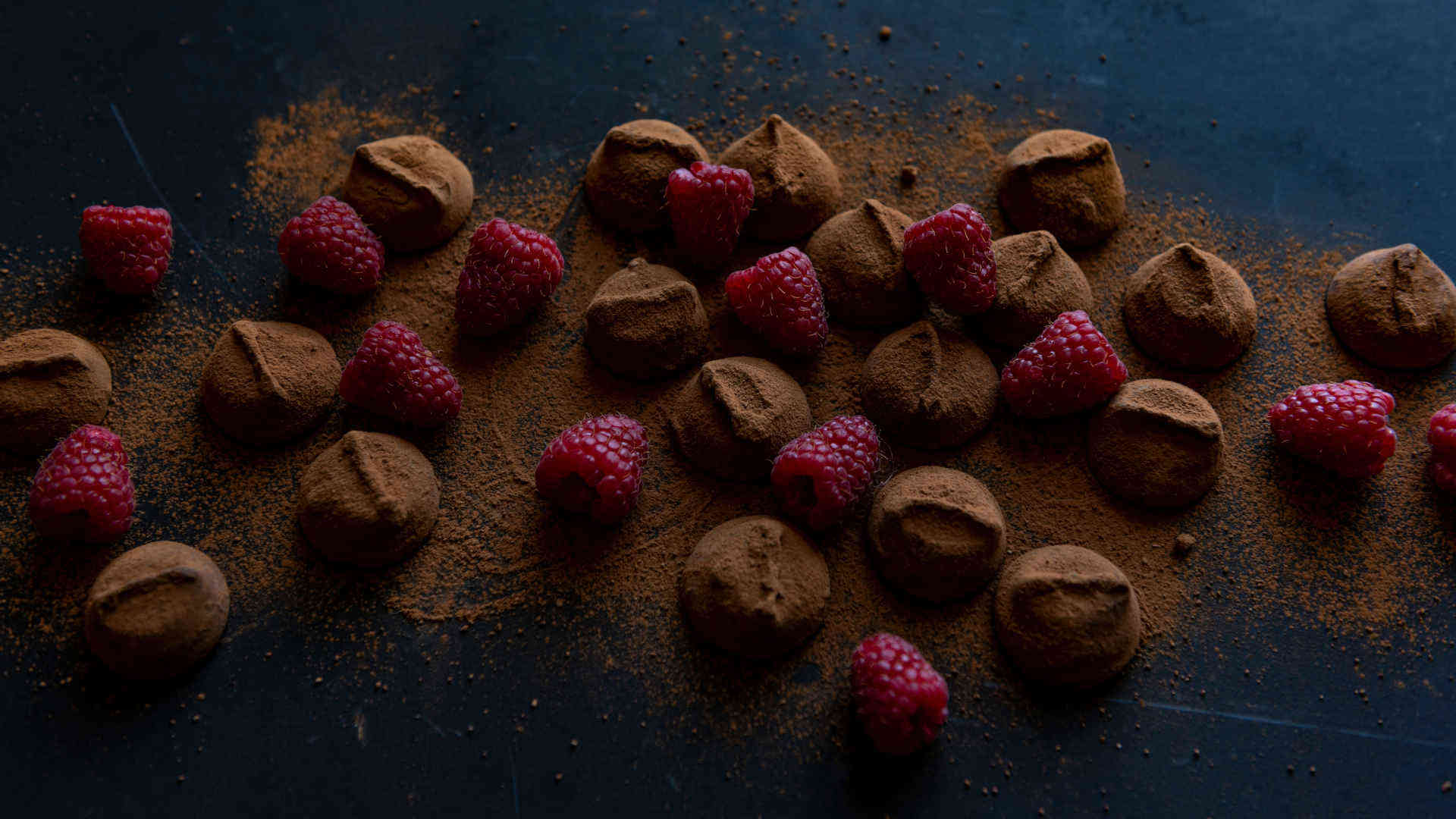 Auf einem blauen Untergrund liegen selbstgemachte, mit Schokoladenpulver überzogene Pralinen und Himbeeren.