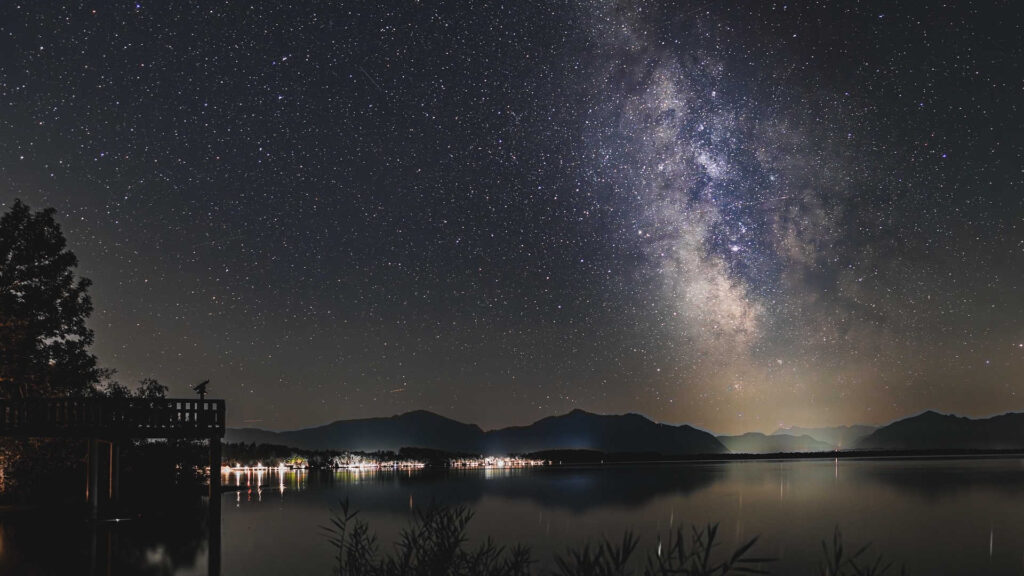 Astrofotografie – So kann man die Milchstraße mithilfe der Fotografie sichtbarmachen. Eine tolle Aufnahme über einem See, mit den Lichtern am Ufer und den Bergen im Hintergrund.