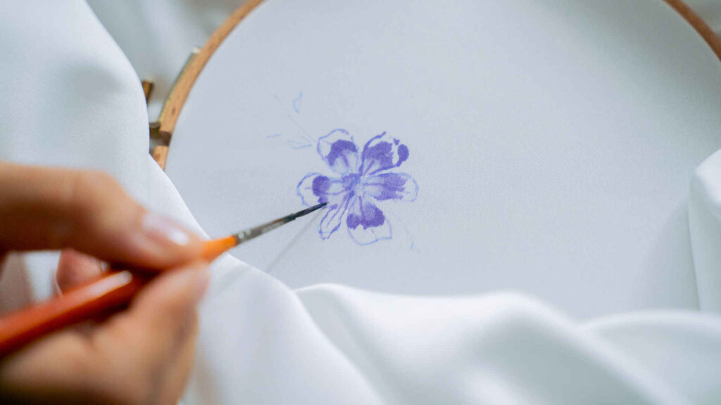 Seidenmalerei - Beginn auf der eingespannten Seide. Vorsichtig wird eine blaue Blume mit dem Pinsel gemalt.